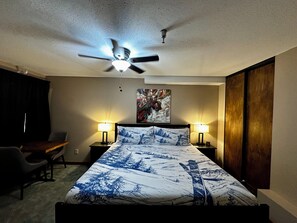 A hotel room with a bed, two bedside lamps, a ceiling fan, a painting on the wall, and a desk with a chair.