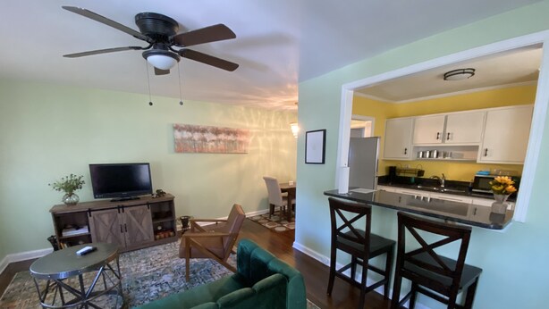 A living room with a green sofa, a TV, and a ceiling fan. There is a kitchen with white cabinets and a dining area with two chairs. - Cozy Atlanta Apt Near Downtown & Airport (Atlanta)