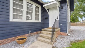 Ein Haus mit einer Veranda, einer Treppe aus Beton, die zu einer Tür führt, und einem kleinen Garten mit einem Blumentopf.