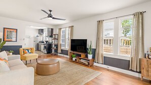 Ein modernes Wohnzimmer mit Sofa, Hocker und TV-Standfuß, mit einer Küche im Hintergrund und großen Fenstern mit Vorhängen.