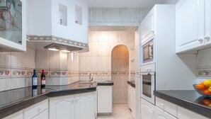 A modern kitchen with white cabinets, a black countertop, and a built-in oven. There are two wine bottles on the counter and a fruit bowl on the right. - Town house for 4/6 people (Torviscas Bajo)