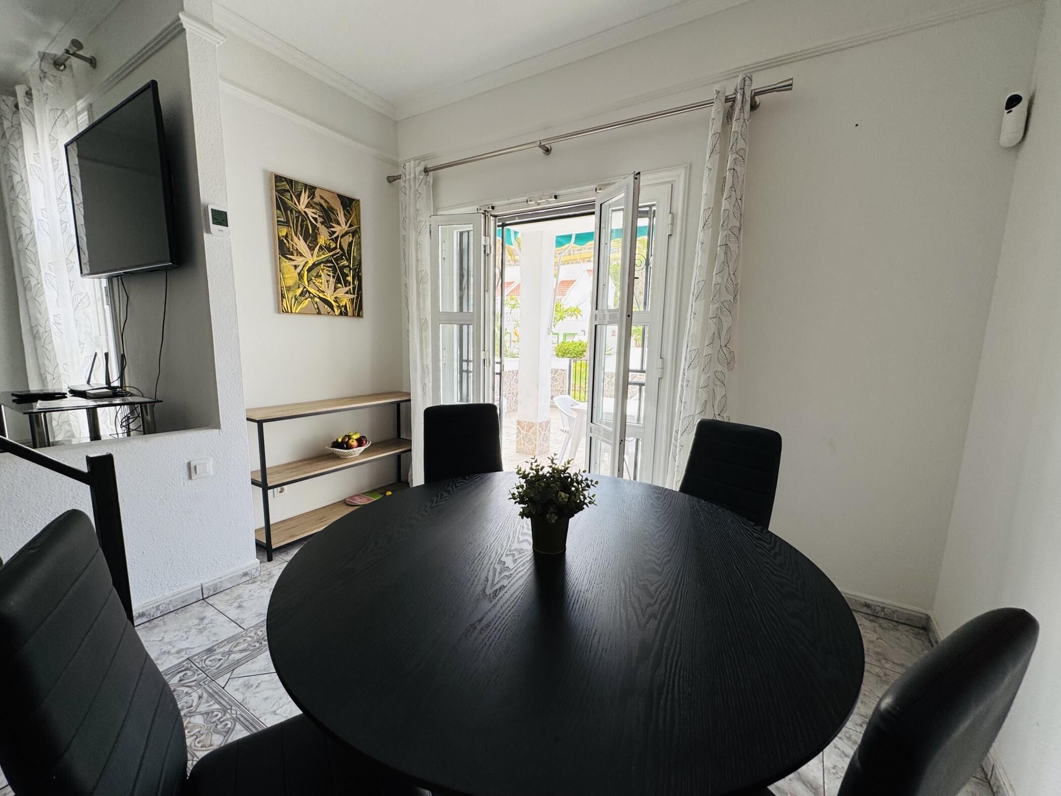 Un comedor con una mesa redonda, sillas negras, una planta pequeña, un televisor en la pared y una ventana con cortinas.
