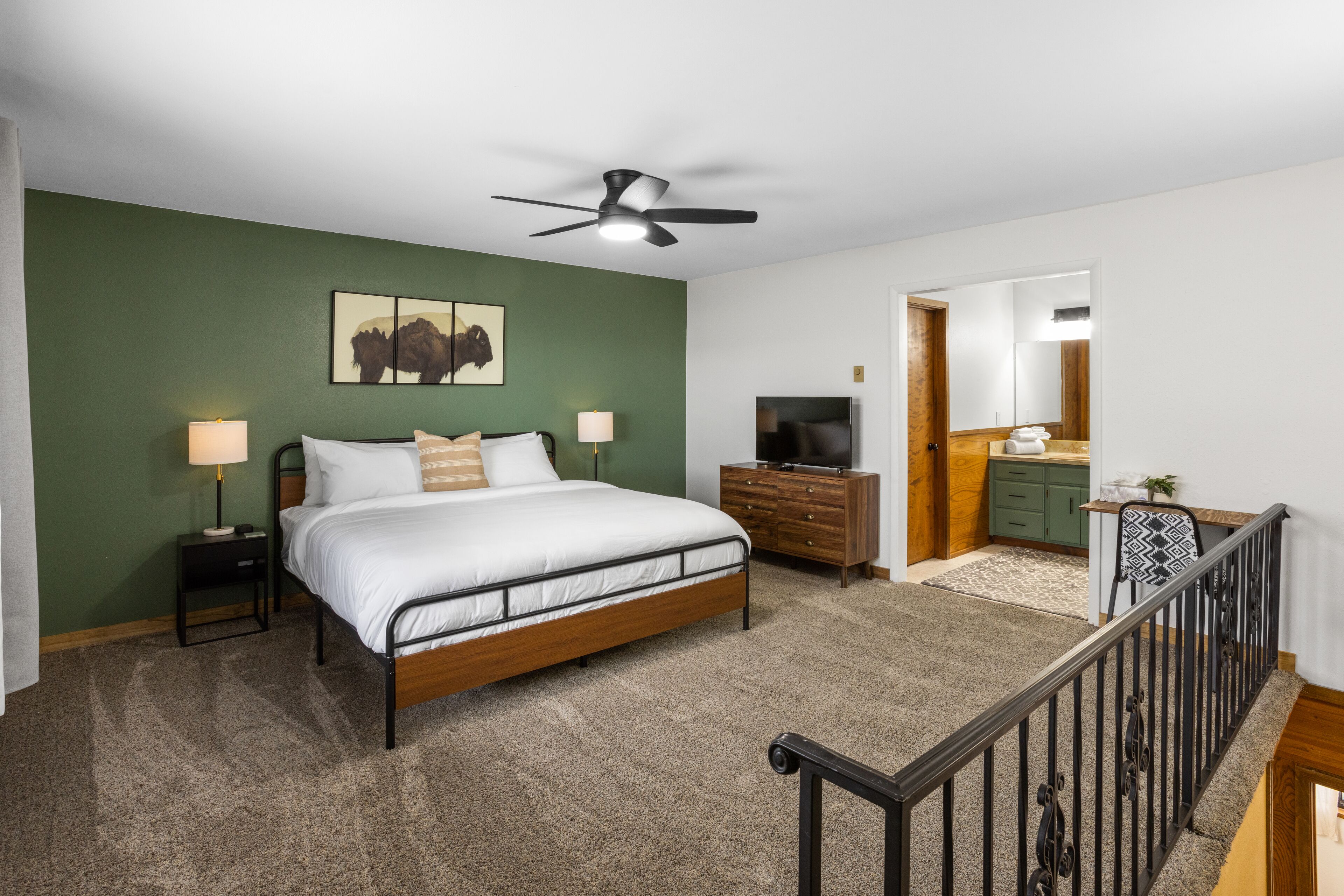 A bedroom with a bed, bedside tables, a ceiling fan, a TV, a dresser, and a bathroom visible through an open door.