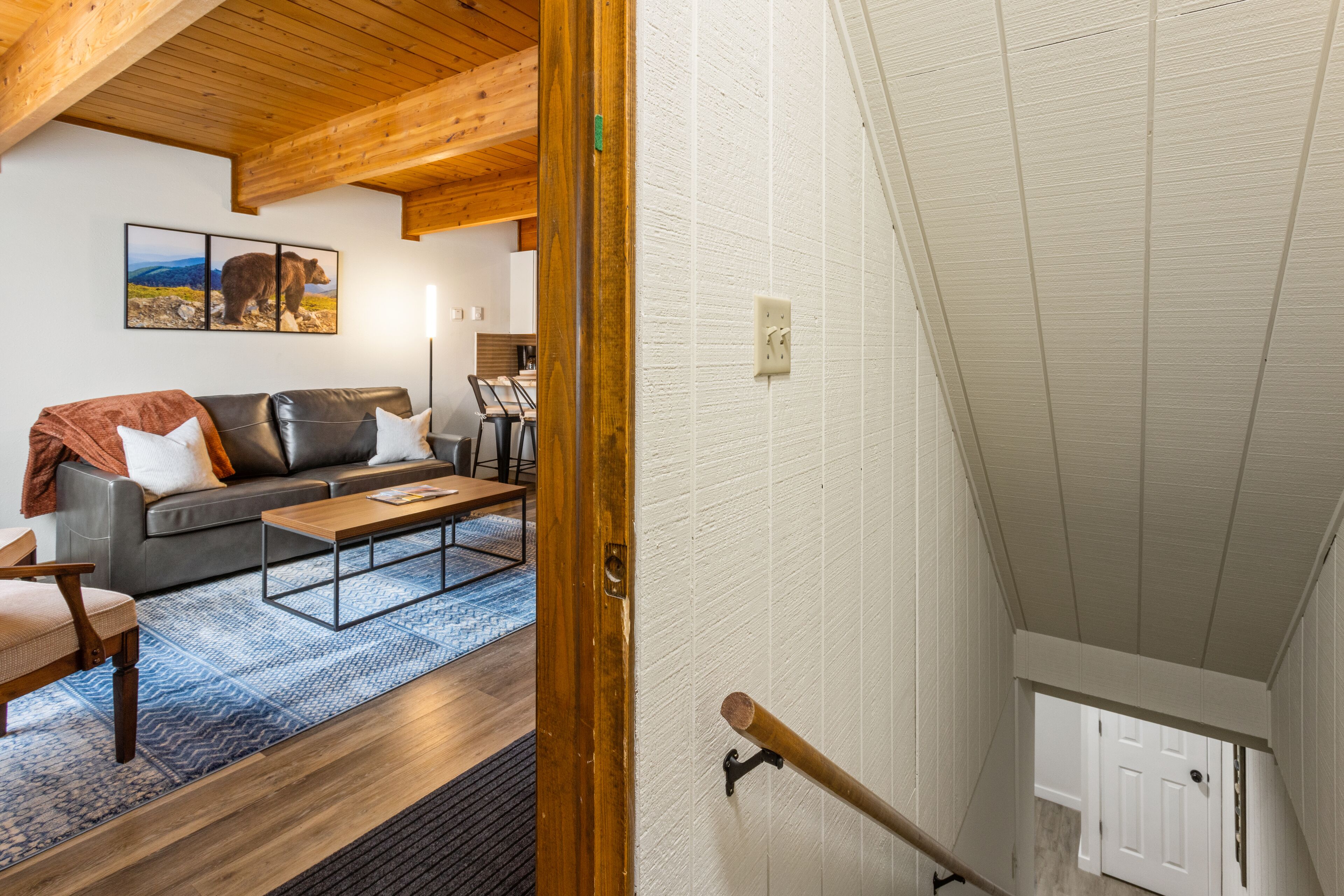 Una sala de estar acogedora con un sofá de cuero, una mesa de centro de madera y un tapete azul estampado. Una escalera de madera lleva a un nivel superior.