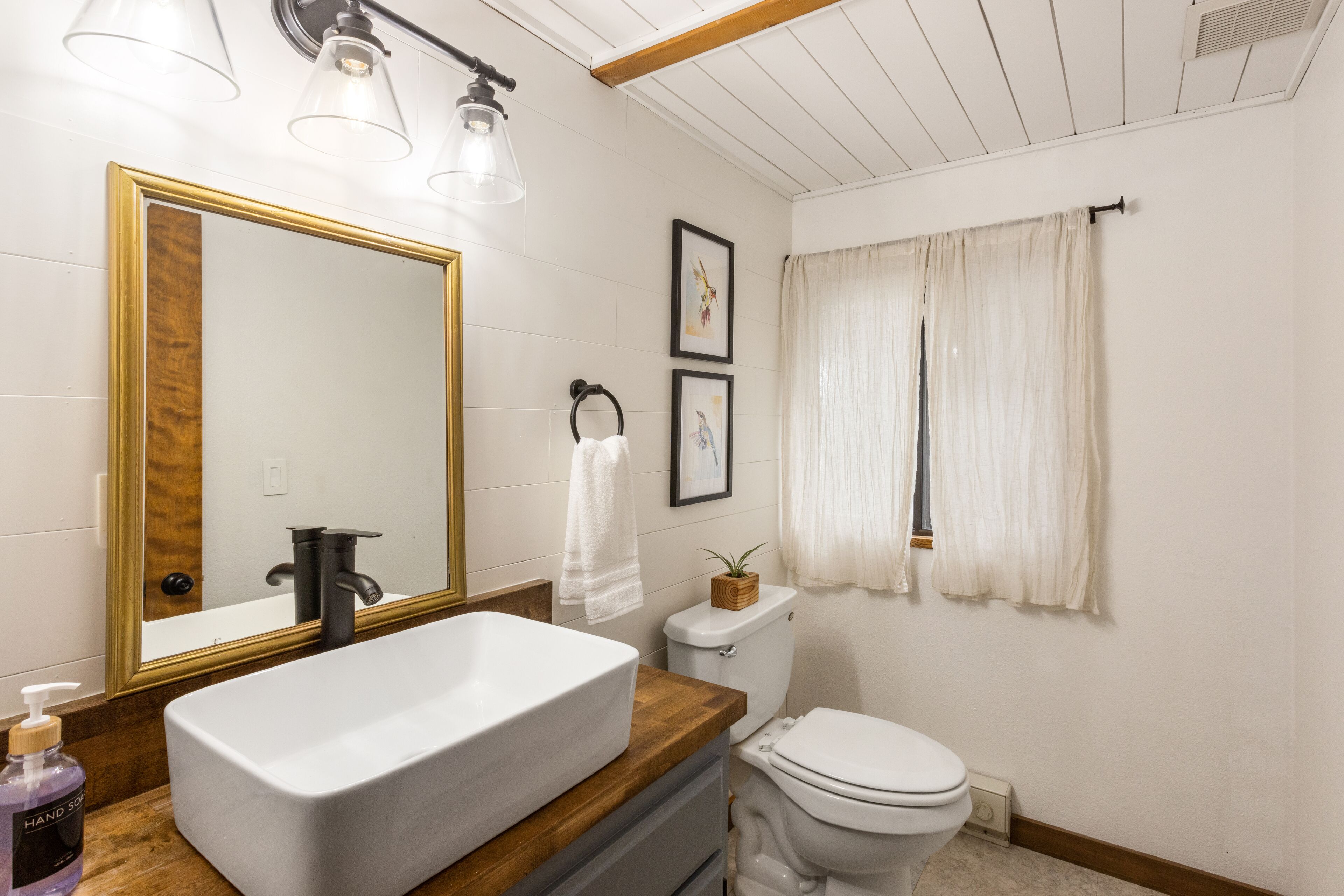 A bathroom with a large mirror, a white sink, a toilet, and a window with curtains.