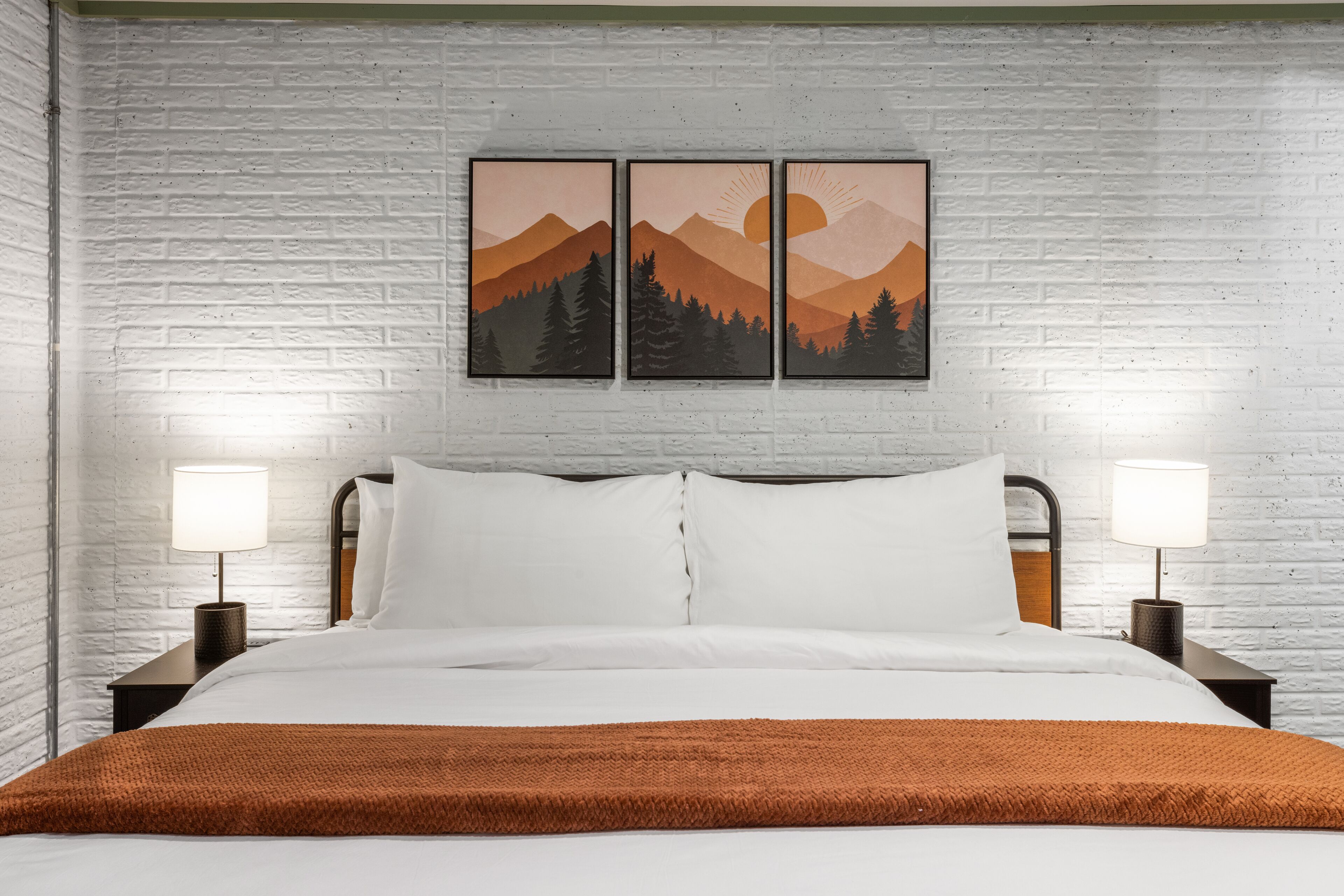 A hotel room with a white brick wall, a bed with white bedding, two bedside lamps, and a brown throw blanket.