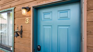 A blue door with a number "3" above it, a window with a curtain, and a wall-mounted light fixture. - Otters Den (West Yellowstone)