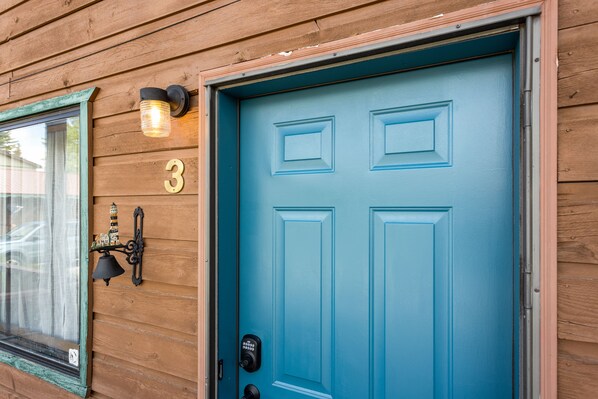 Eine blaue Tür mit der Zahl „3“ darüber, ein Fenster mit Vorhang und eine Wandleuchte.