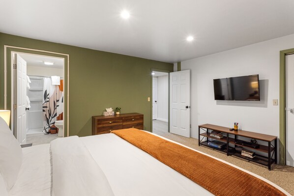 A bedroom with a bed, a TV mounted on the wall, a wooden dresser, and a console table with books. - Otters Den (West Yellowstone)