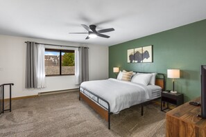Ein Schlafzimmer mit einem Bett, einem Deckenventilator, einem Nachttisch mit Lampe, einem Fernseher und einem Fenster mit Vorhängen.