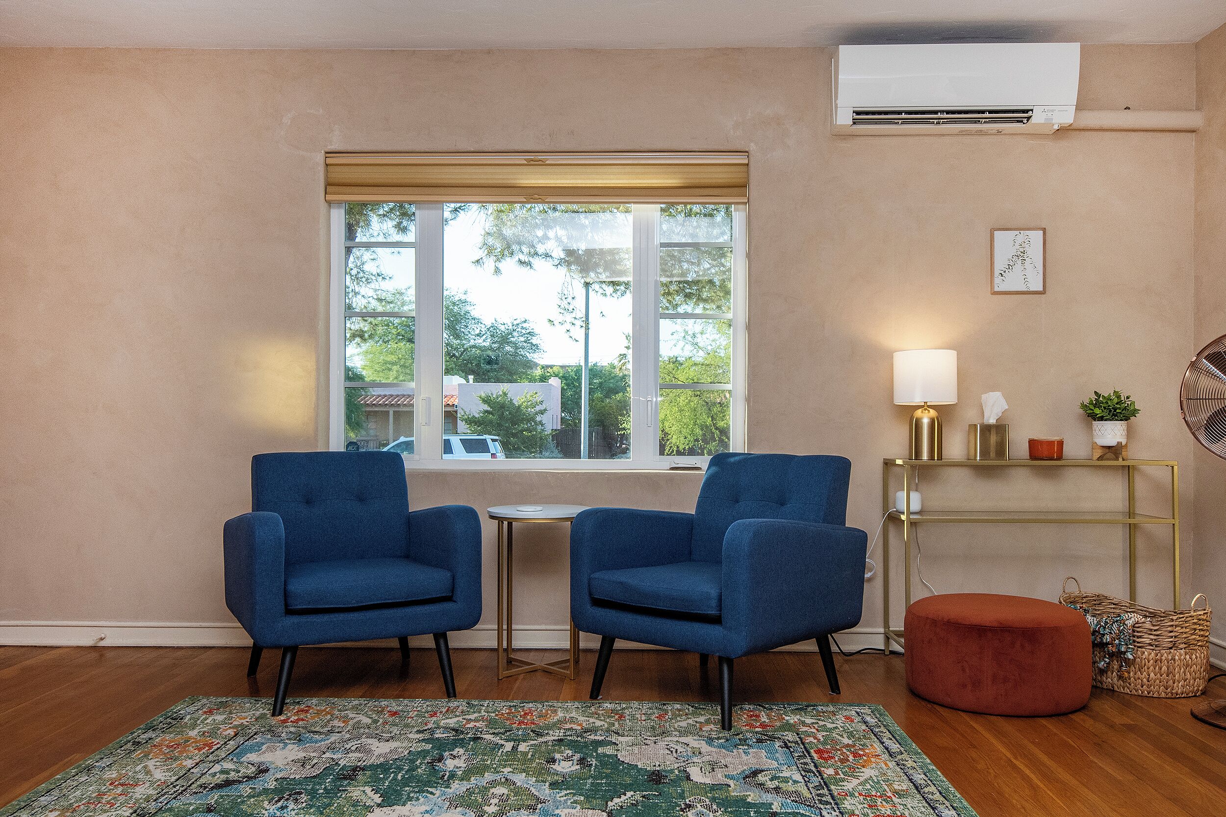 Una habitación con dos sillones azules, una mesita pequeña, un reposapiés rojo, un tapete estampado, una consola dorada con una lámpara y un equipo de aire acondicionado.