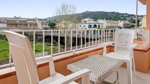 Un balcon avec des chaises blanches et une table en osier, donnant sur un quartier résidentiel avec des maisons et une colline en arrière-plan.