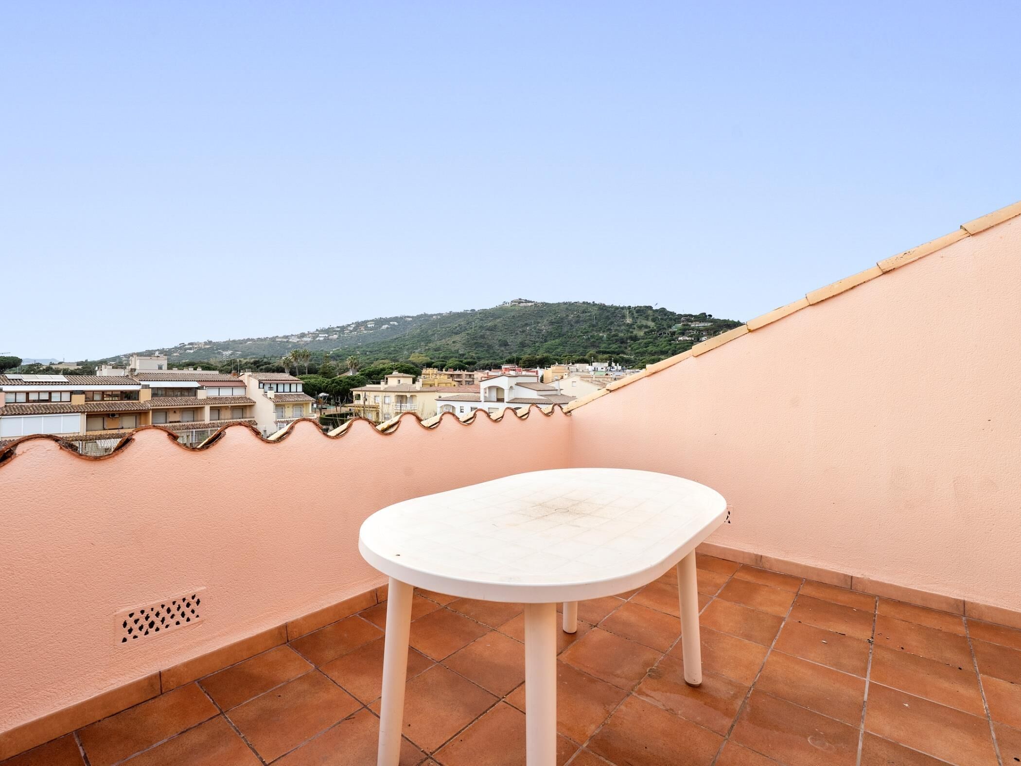 A rooftop terrace with a white table and chairs, terracotta tiles, and a view of a hillside town.