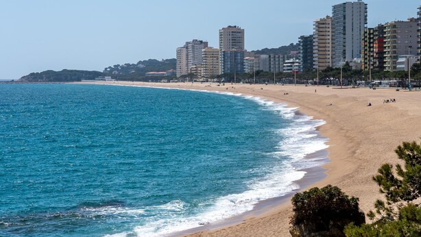 Une plage côtière avec un rivage sablonneux et une mer bleue et limpide. Au loin, on aperçoit plusieurs immeubles de grande hauteur.