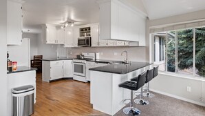 A modern kitchen with white cabinetry, a central island with bar stools, a microwave, oven, and a large window offering a view of trees. - Spacious Mountain Hideaway 4 BD 3 BA W/ Hot Tub (Ogden)