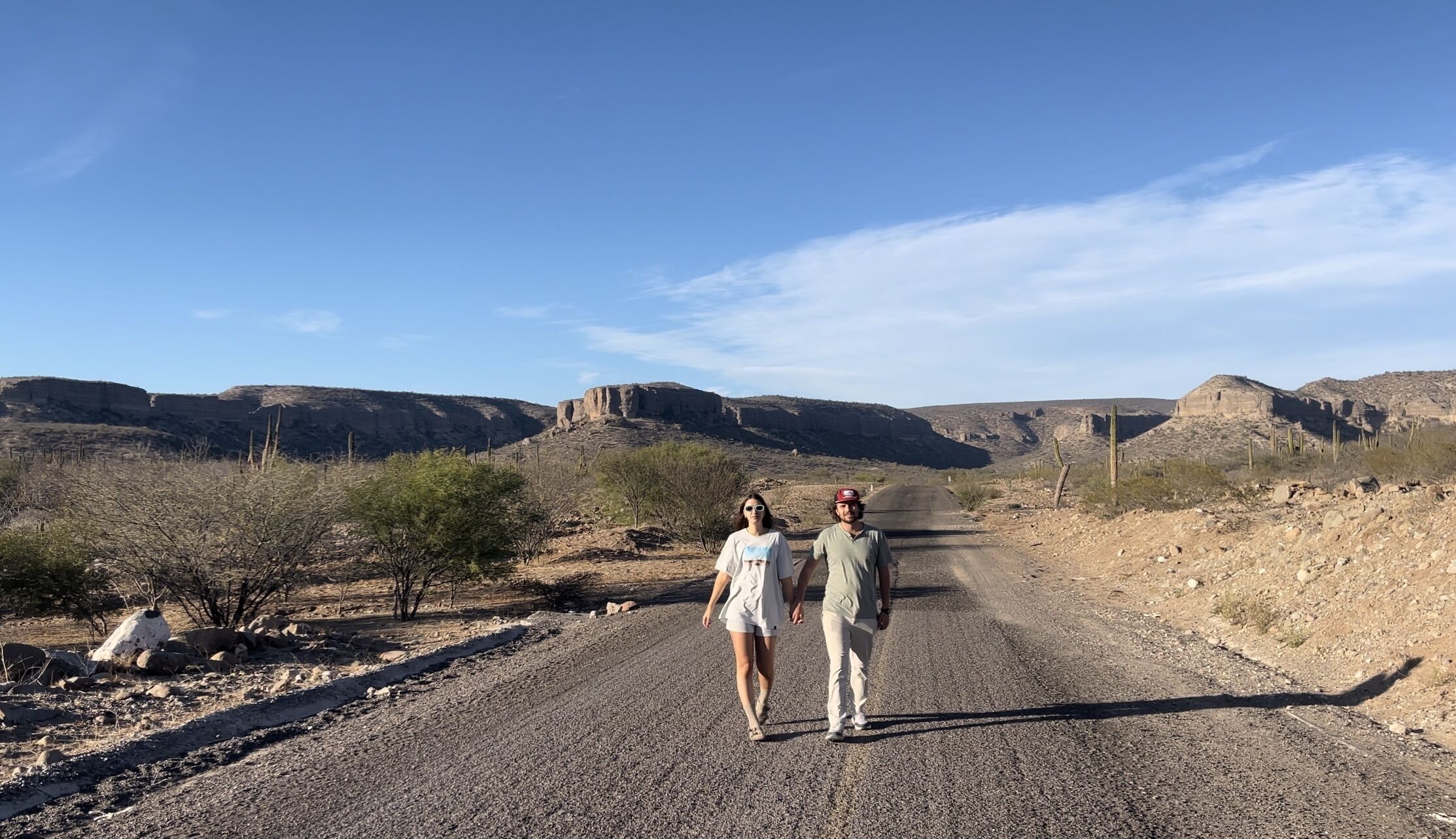 Un couple se promène sur un chemin de gravier, dans un paysage désertique, avec des collines rocheuses en arrière-plan.