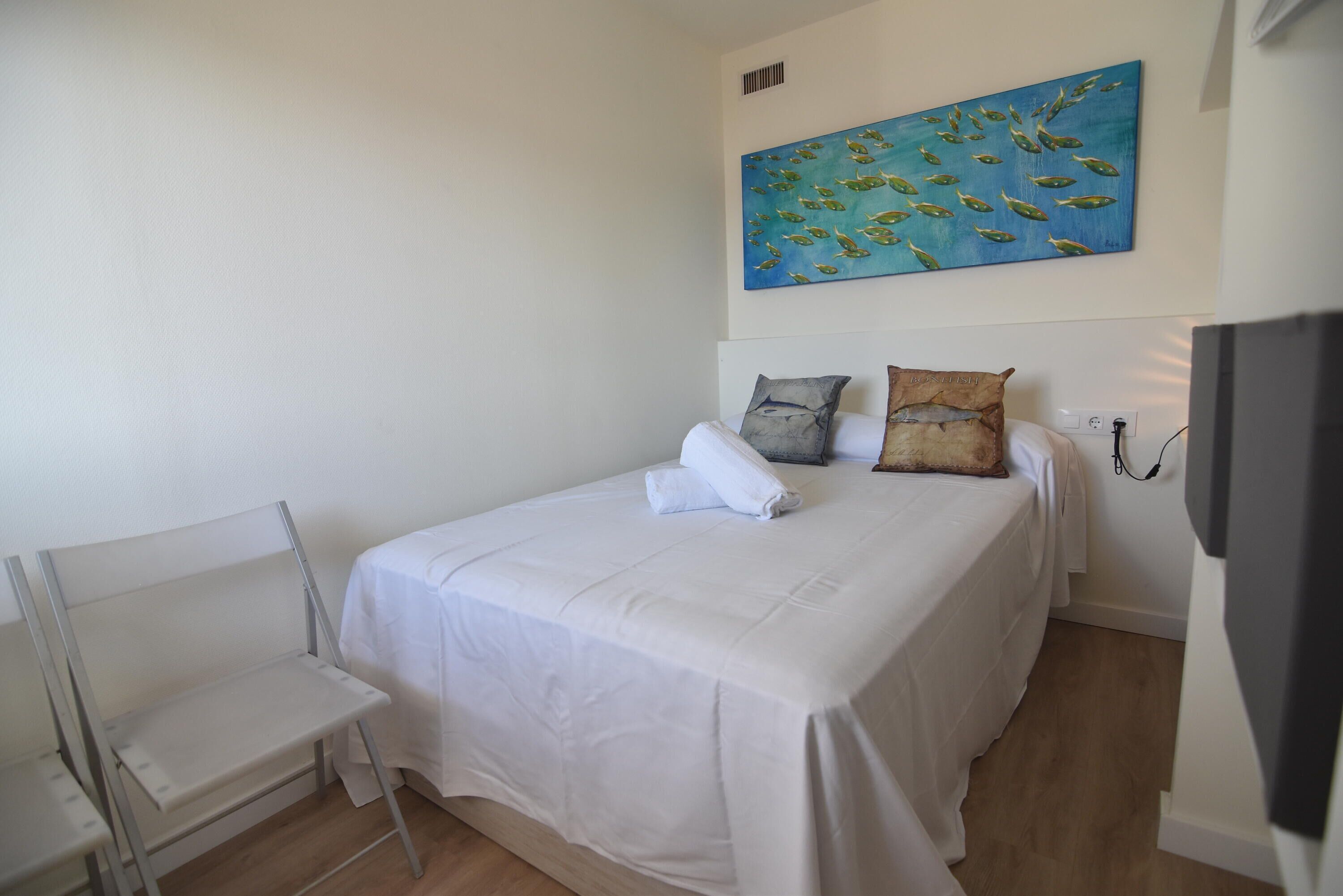 A neatly made bed with white linens, a small bedside table, and a painting of fish on the wall.