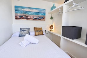 A neatly made bed with white linens, two decorative pillows, a rolled towel, a painting of boats on the wall, a shelf with a blue bottle and a green object, and a white hanging garment rack.