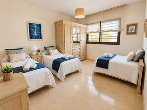 Quarto de hotel com duas camas, um guarda-roupa, uma escrivaninha, uma luminária, uma planta e uma janela com cortinas.