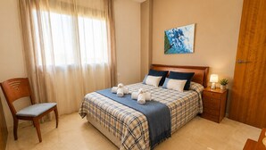 Un dormitorio con cama, una silla, una mesita de noche, una lámpara y un cuadro en la pared.
