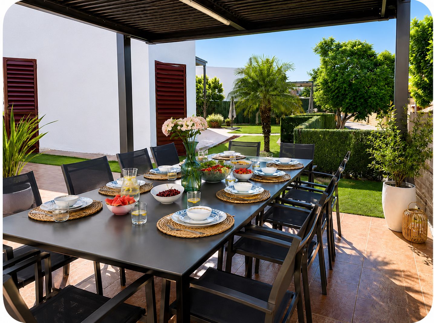 Uma área de refeições ao ar livre com uma mesa comprida posta para uma refeição, rodeada por cadeiras e vista para um jardim bem cuidado.