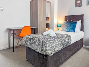 A bedroom with a bed, a desk, an orange chair, a wardrobe, and a wall-mounted lamp.
