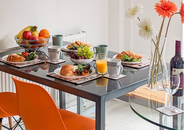 A breakfast table with croissants, fruit, and juice, a vase with flowers, and a wine bottle.