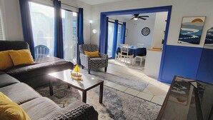 Un salon comprenant un canapé, une table basse et un fauteuil à motifs. On y trouve également un coin repas avec une table et des chaises, ainsi qu’une cuisine équipée de placards et d’un réfrigérateur.