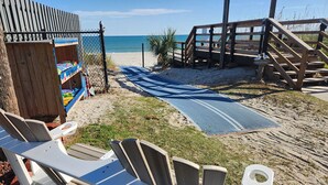 Un espace aménagé au bord de la plage, avec des chaises longues en bois, une structure en bois portant une pancarte « Bienvenue » et un tapis bleu menant à la plage.