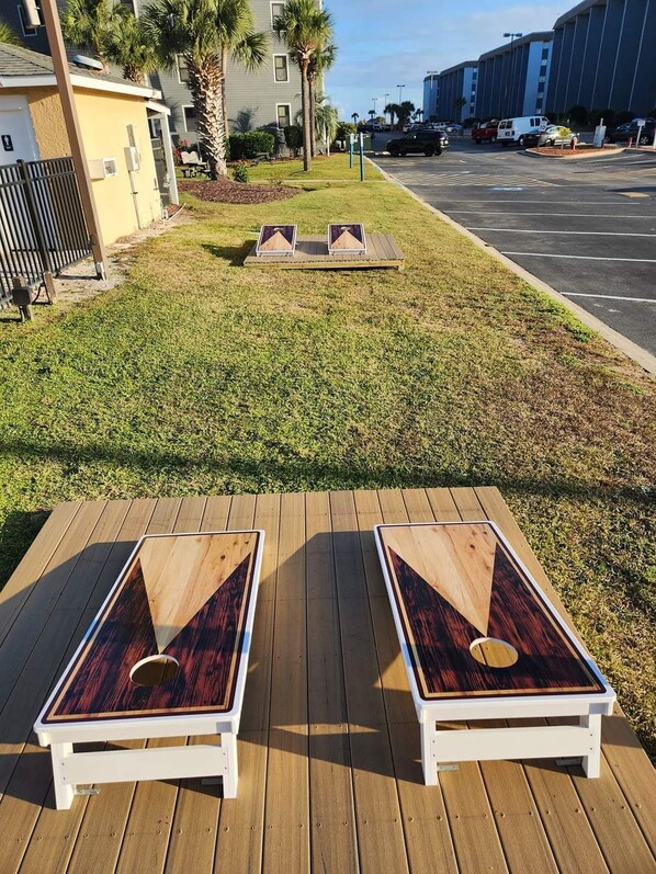 A deck with two lounge chairs and a small wooden table, surrounded by a grassy area with palm trees and a parking lot in the background.