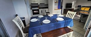 A dining area with a blue tablecloth, white plates, and cutlery. There is a kitchen in the background with white cabinets and a coffee bar.