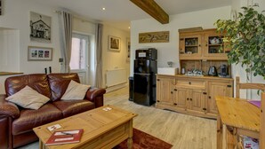 A living room with a leather sofa, wooden coffee table, and a kitchen area with wooden cabinets and a refrigerator. - The Old Post Office, Exford (Exford)