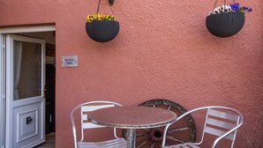 A small outdoor seating area with a round table and two metal chairs, a rustic wooden wheel, and two hanging flower baskets against a pink wall. - The Old Post Office, Exford (Exford)