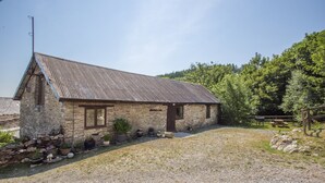 Un bâtiment rustique en pierre, doté d’un toit métallique incliné, entouré d’une allée en gravier et de plantes en pot.