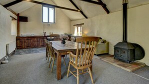 Un grand espace à manger avec une table et des chaises en bois, une cuisine à l’arrière-plan et un poêle à bois.