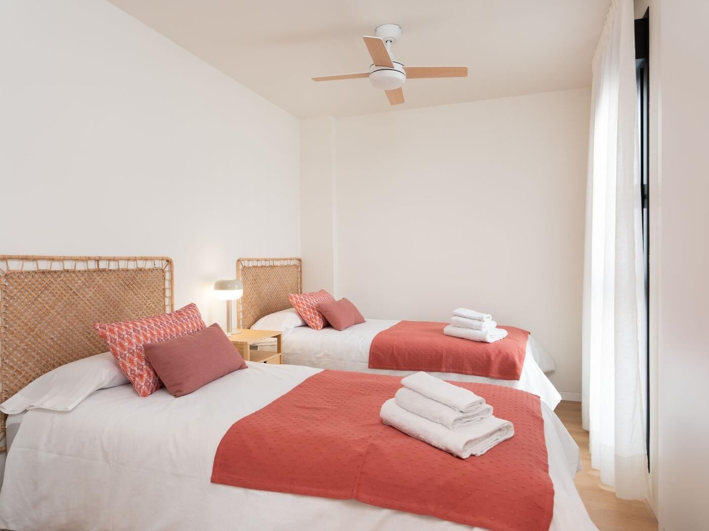 Ein Hotelzimmer mit zwei Betten, jeweils mit roter Decke und Kissen, einem Deckenventilator und einem Nachttisch mit Lampe.