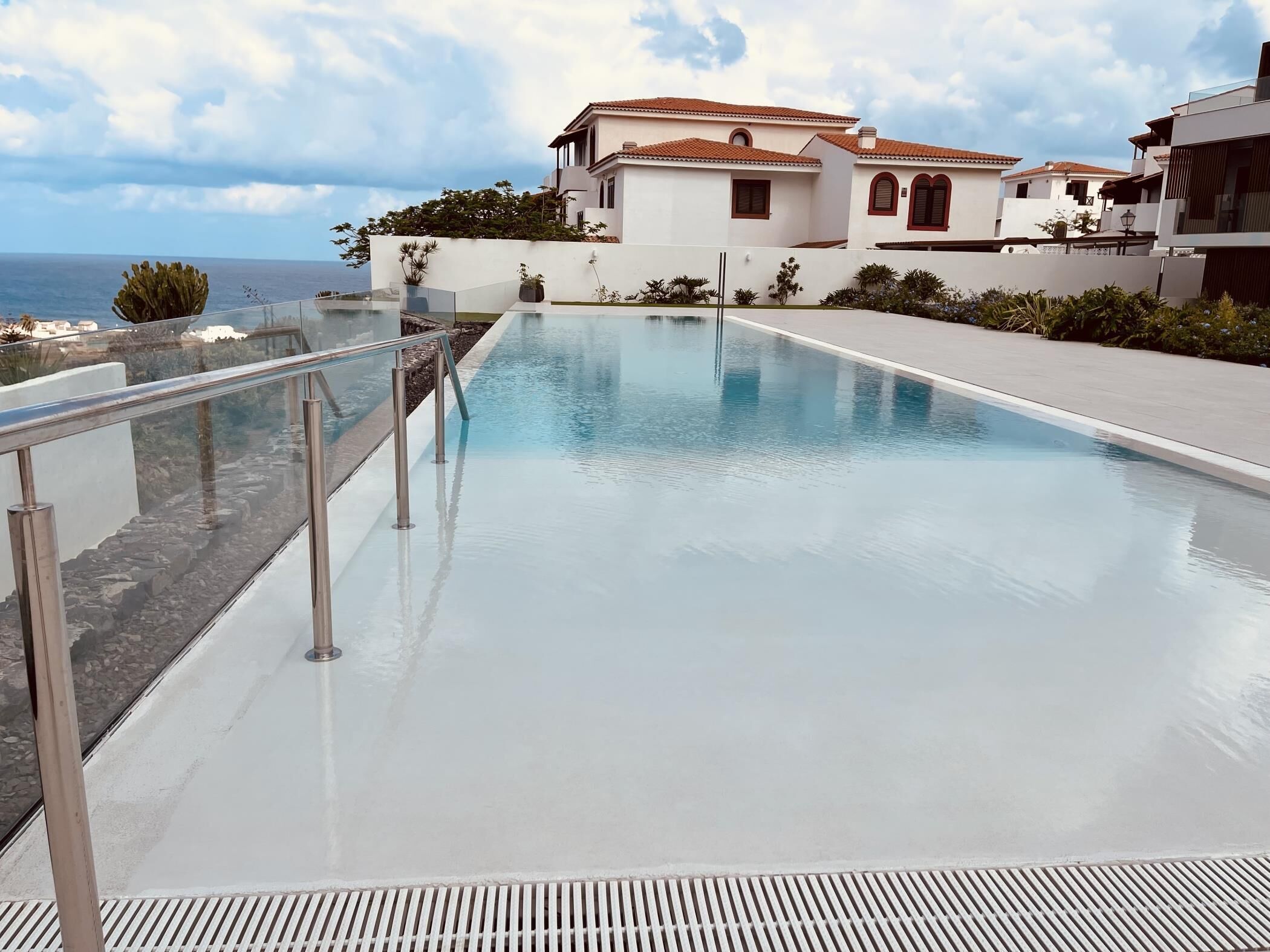 Ein Schwimmbecken mit Glastrennwand, ein weißes Gebäude mit roten Ziegeldächern und Blick auf den Ozean.