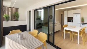 Una cocina moderna con armarios blancos, un microondas y un refrigerador. Hay un comedor con una mesa y sillas, y una puerta corrediza de vidrio que da a un balcón con plantas.