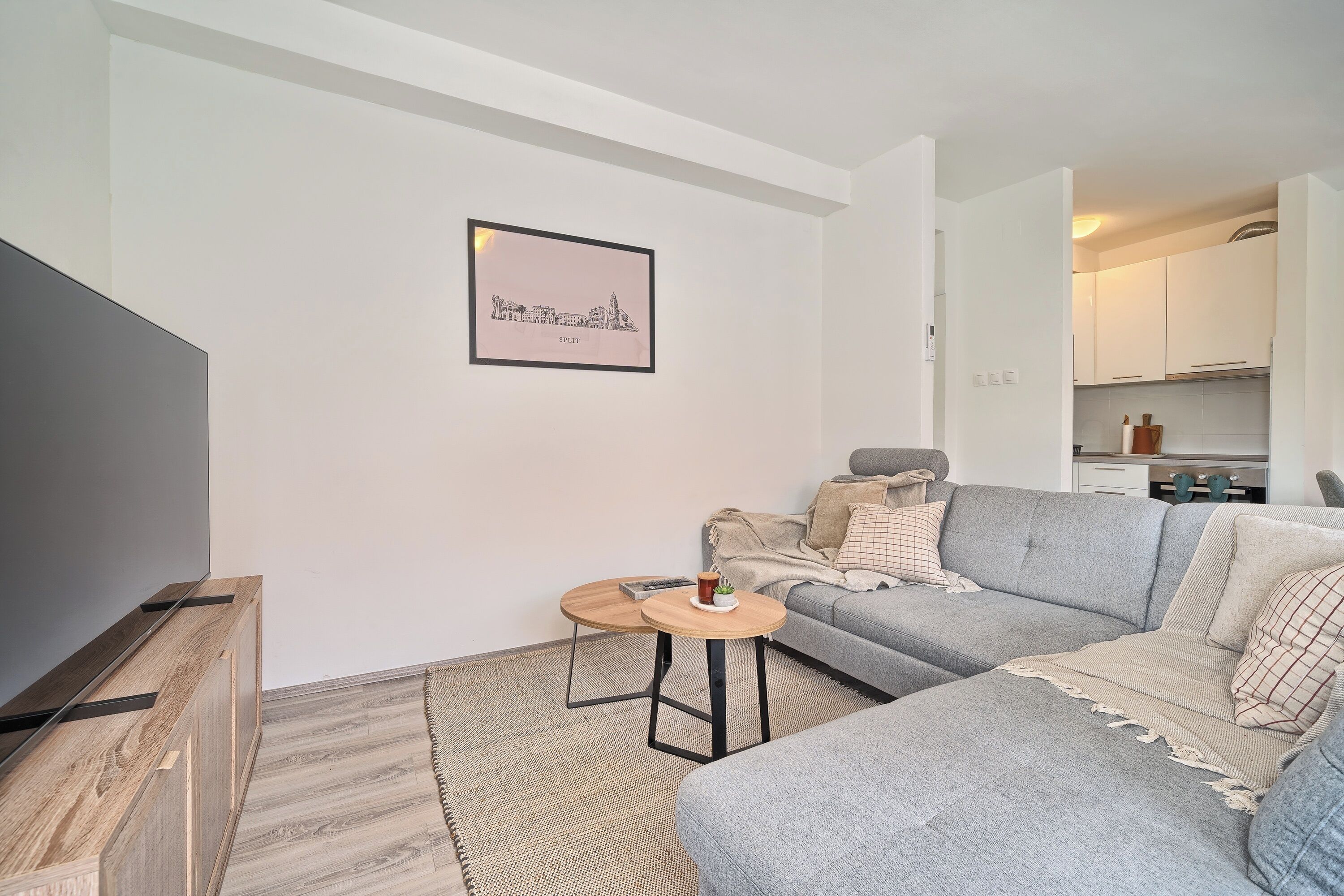 A modern living room with a grey sofa, a small round wooden table, a flat-screen TV, and a framed cityscape picture on the wall.