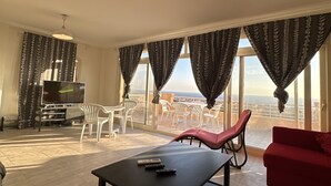 A living room with a TV, a red sofa, a black coffee table, and a balcony with white chairs and a red lounge chair.
