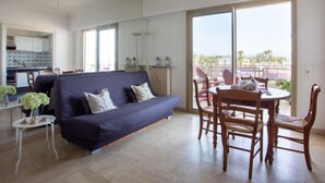 A modern living room with a sofa, dining table, and chairs. There is a kitchen area with cabinets and a window. - le corail by Interhome (Villeneuve-Loubet)