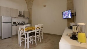 Una cocina compacta con armarios blancos, un horno empotrado, un microondas y un comedor con sillas y mesa blancos. Un televisor está montado en la pared, y una licuadora amarilla sobre la encimera.