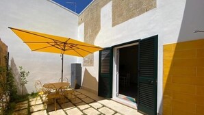 Un patio con mesa y sillas bajo un paraguas amarillo, una puerta con contraventanas verdes y una pared blanca con sección de ladrillo.
