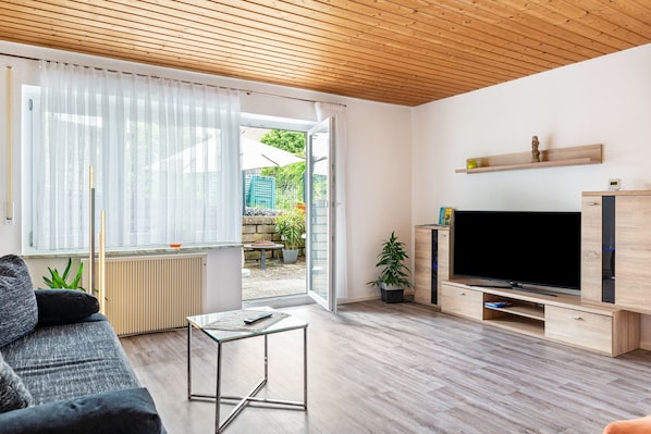 A living room with a sofa, a coffee table, a TV, and a sliding glass door leading to an outdoor area with plants.