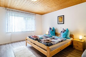 A wooden bed with a quilt, a bedside table with a lamp, and a window with sheer curtains.