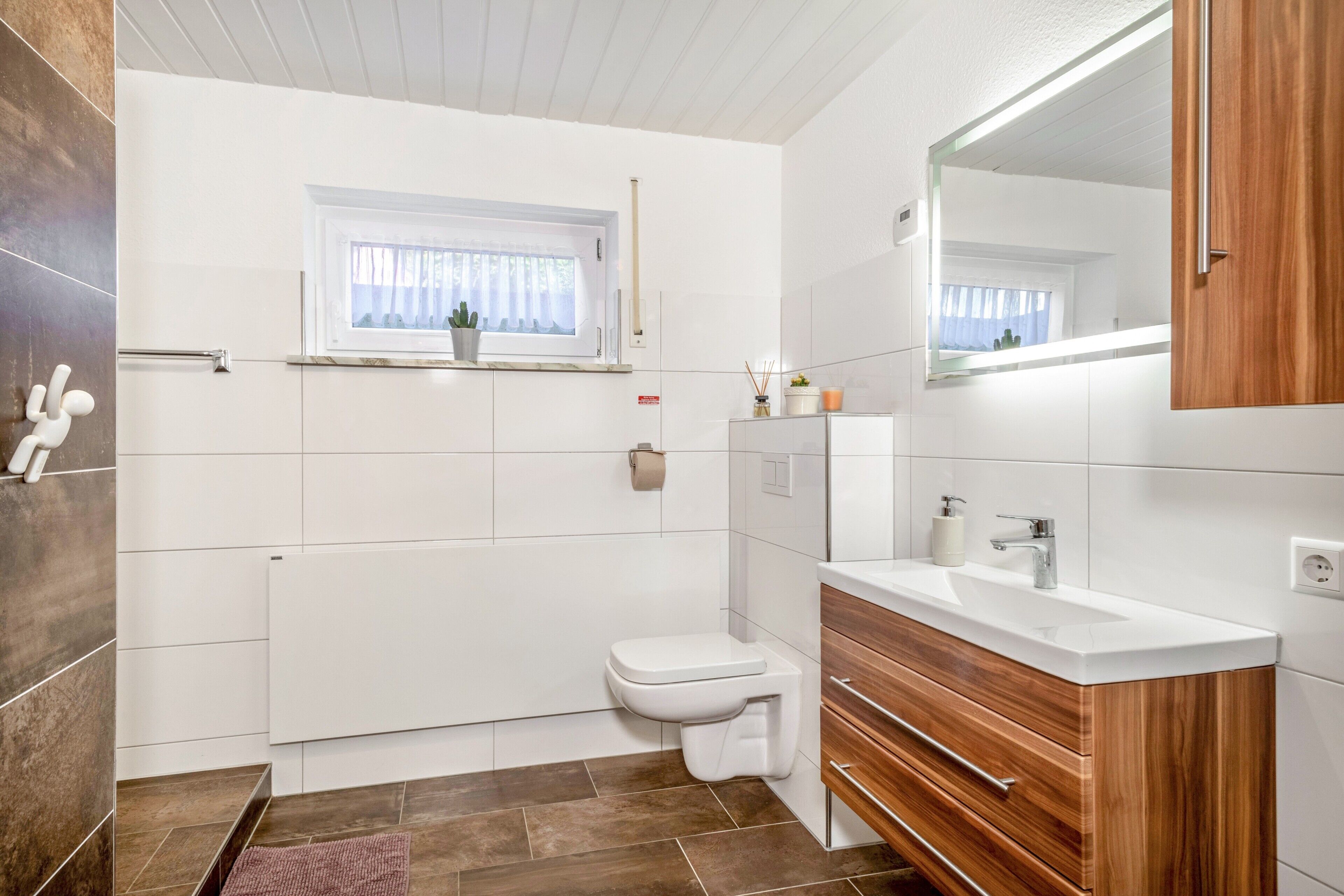 A modern bathroom with a white toilet, wooden vanity, and a window with a plant.
