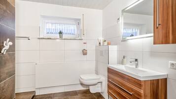 A modern bathroom with a white toilet, wooden vanity, and a window with a plant.