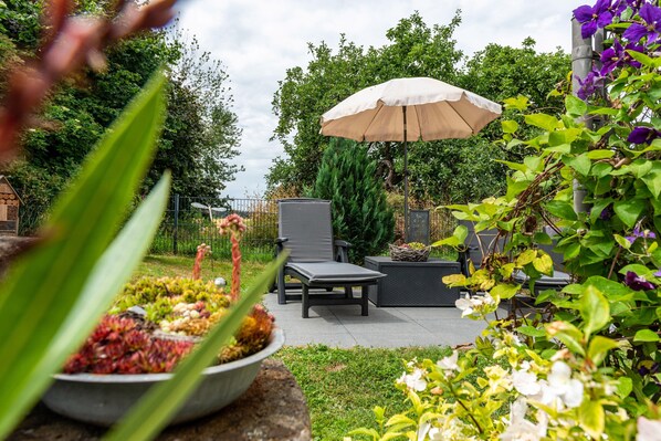 An outdoor seating area with chairs and a table, surrounded by plants and a pergola with an umbrella.