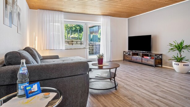 A modern living room with a grey sofa, a glass coffee table, a TV, and a wooden ceiling.