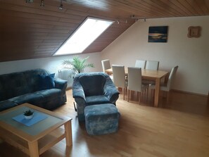 A loft-style living space with a sloped ceiling, a dining area with a wooden table and chairs, a sofa, a chair, and a small table with a plant.